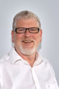 Älterer Mann mit weißen Haaren, Brille und weißem Hemd lächelt vor hellem Hintergrund.