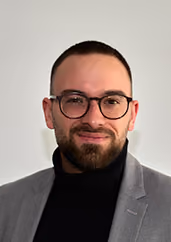 Mann mit Brille und kurzem Bart, trägt schwarzen Rollkragenpullover und graues Sakko vor weißem Hintergrund.