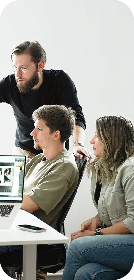 Three people collaborating in a bright office, two seated and one standing, focusing on a laptop screen.