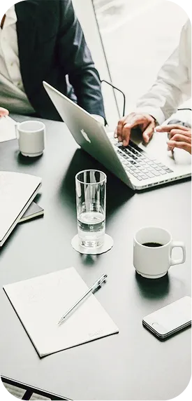 Two people working on laptops at a desk with a glass of water, coffee cup, notebook, pen, and smartphone.