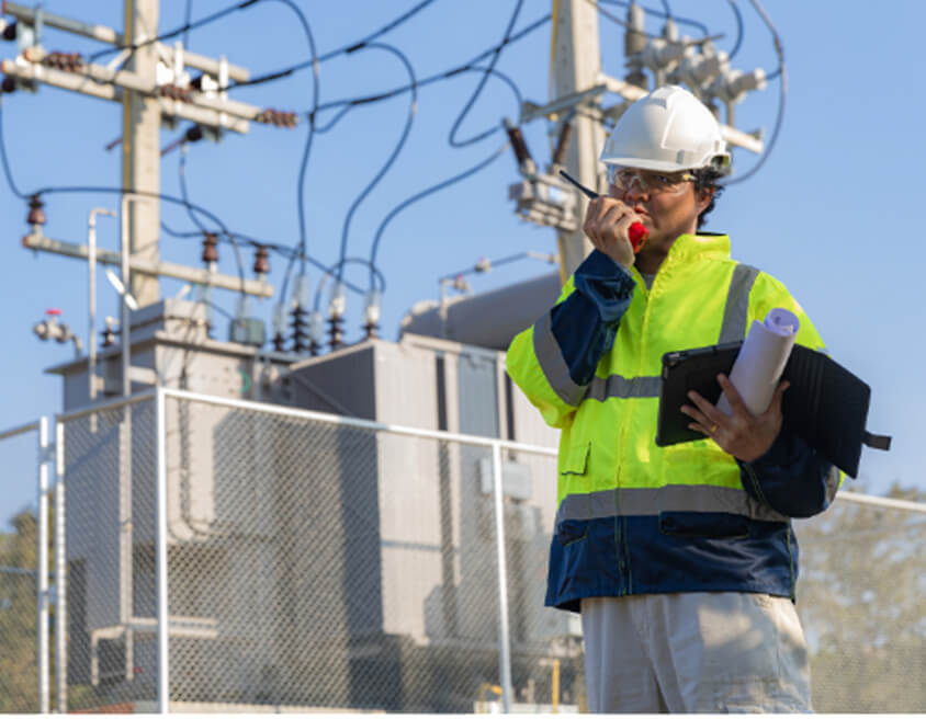 A utility worker, wearing a hard hat and high-visibility jacket, is speaking into a walkie-talkie near a power substation surrounded by electrical equipment and transmission lines.