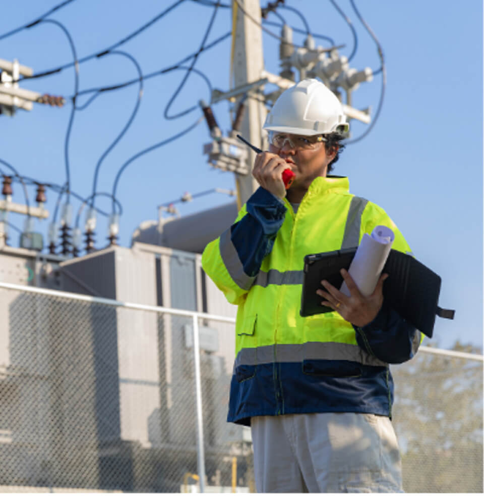 A utility worker, wearing a hard hat and high-visibility jacket, is speaking into a walkie-talkie near a power substation surrounded by electrical equipment and transmission lines.