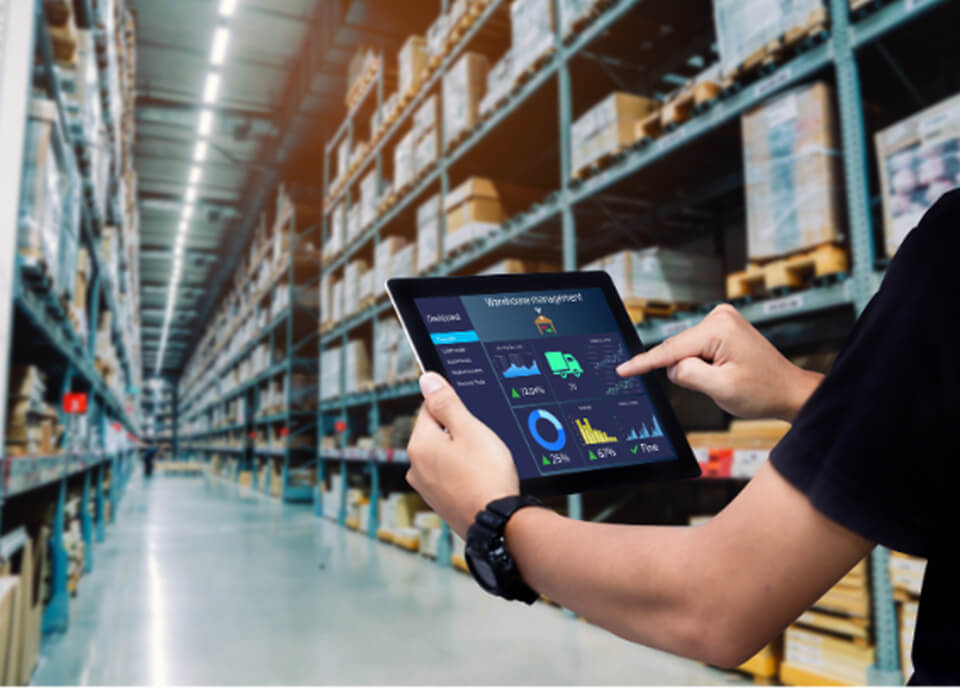 A person holding a tablet in a warehouse, displaying a logistics dashboard with graphs and statistics. Shelves filled with boxes and pallets are visible in the background, indicating a busy storage environment.