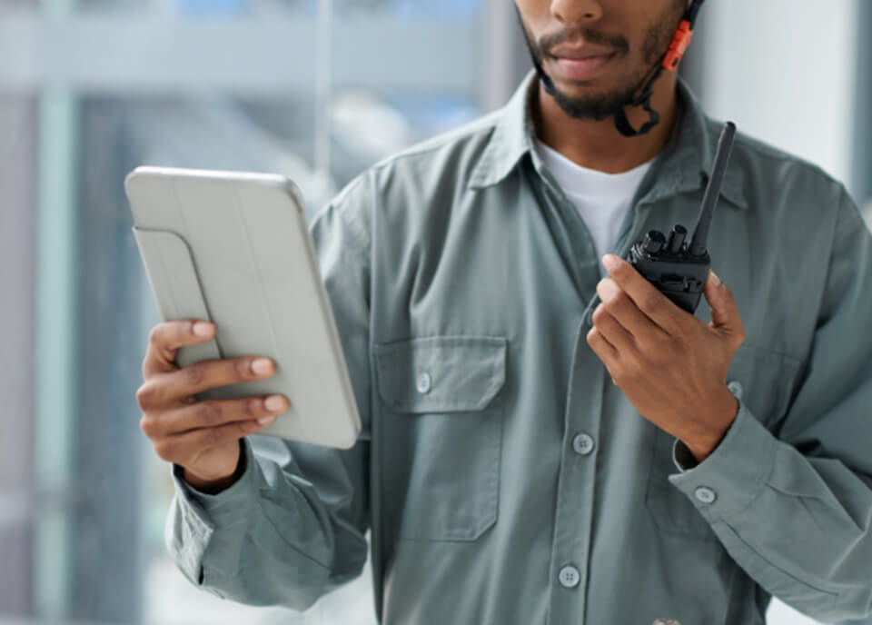 A person wearing a helmet and a gray shirt holds a tablet in one hand and a walkie-talkie in the other, appearing engaged in communication or monitoring an activity in an indoor setting.