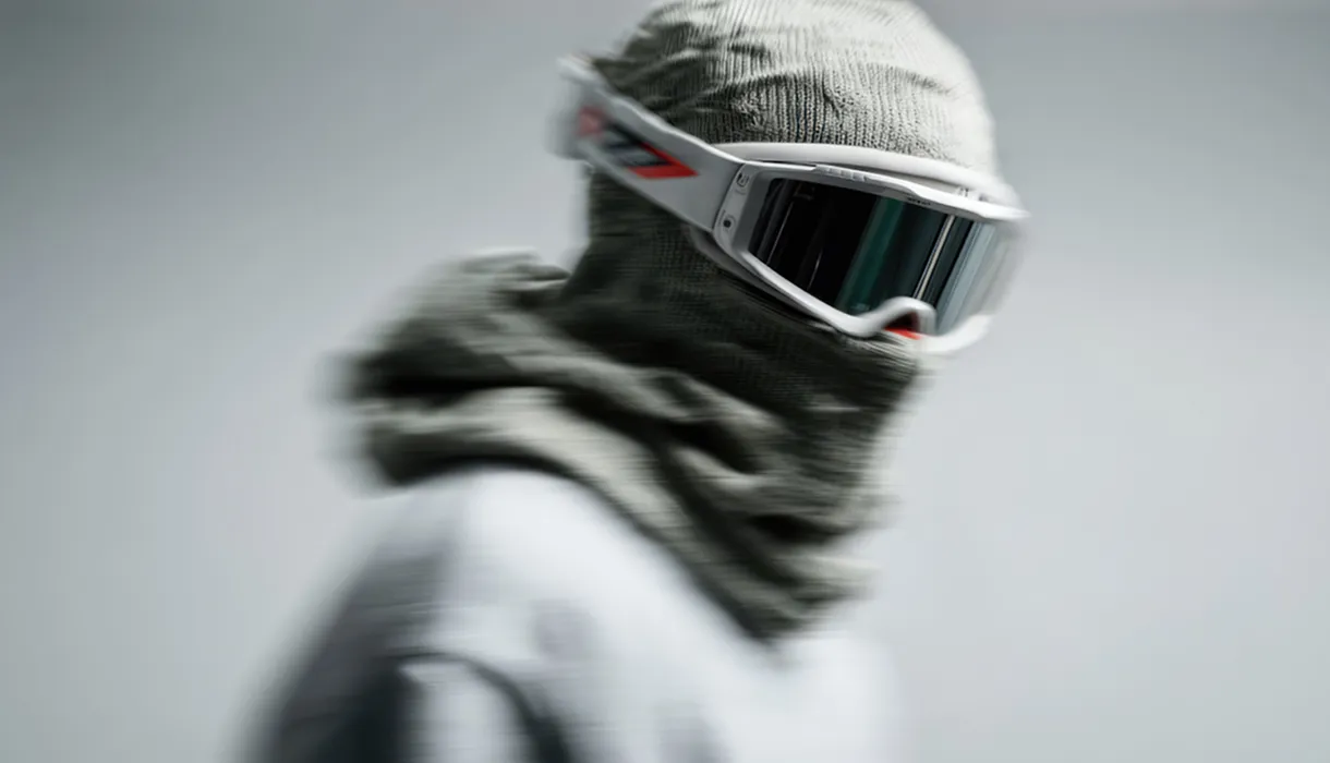 Person wearing a gray balaclava and white ski goggles with a blurred motion effect on a gray background.