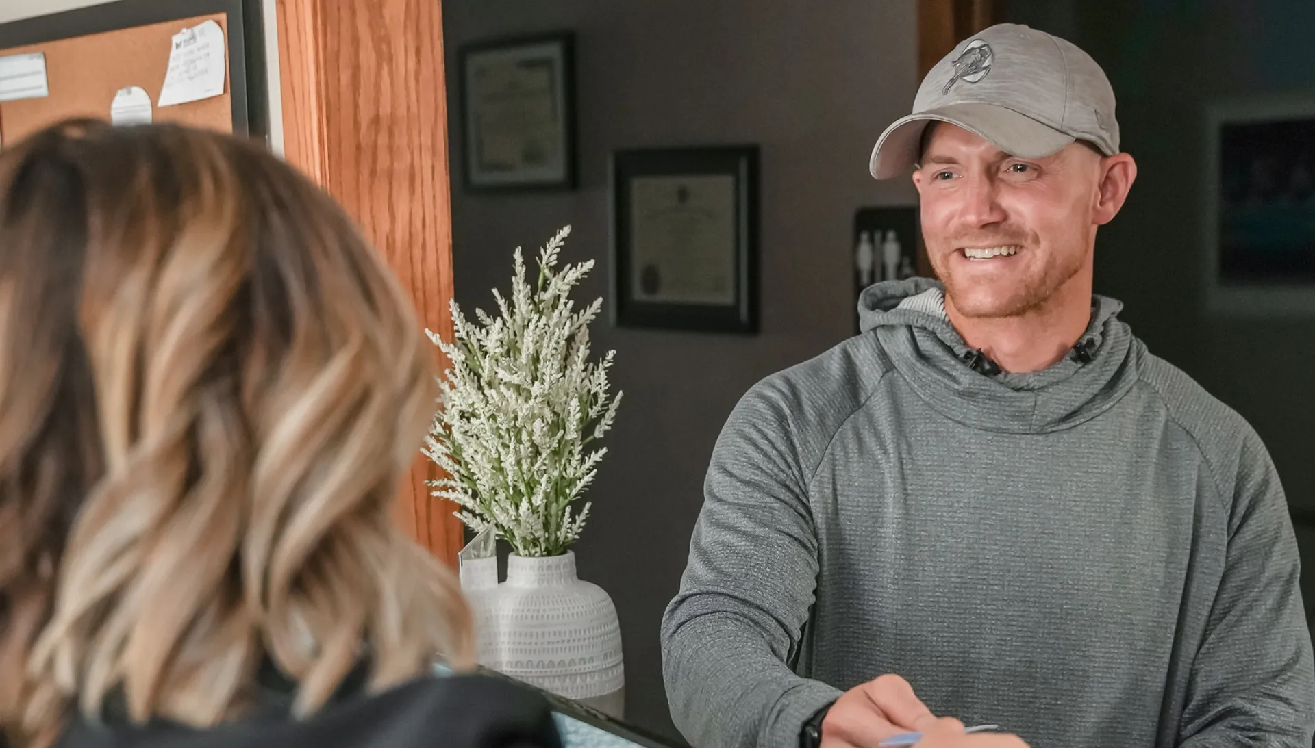 Smiling man in a gray hoodie and cap handing a card to a receptionist at a desk with a white vase and plant in the background.