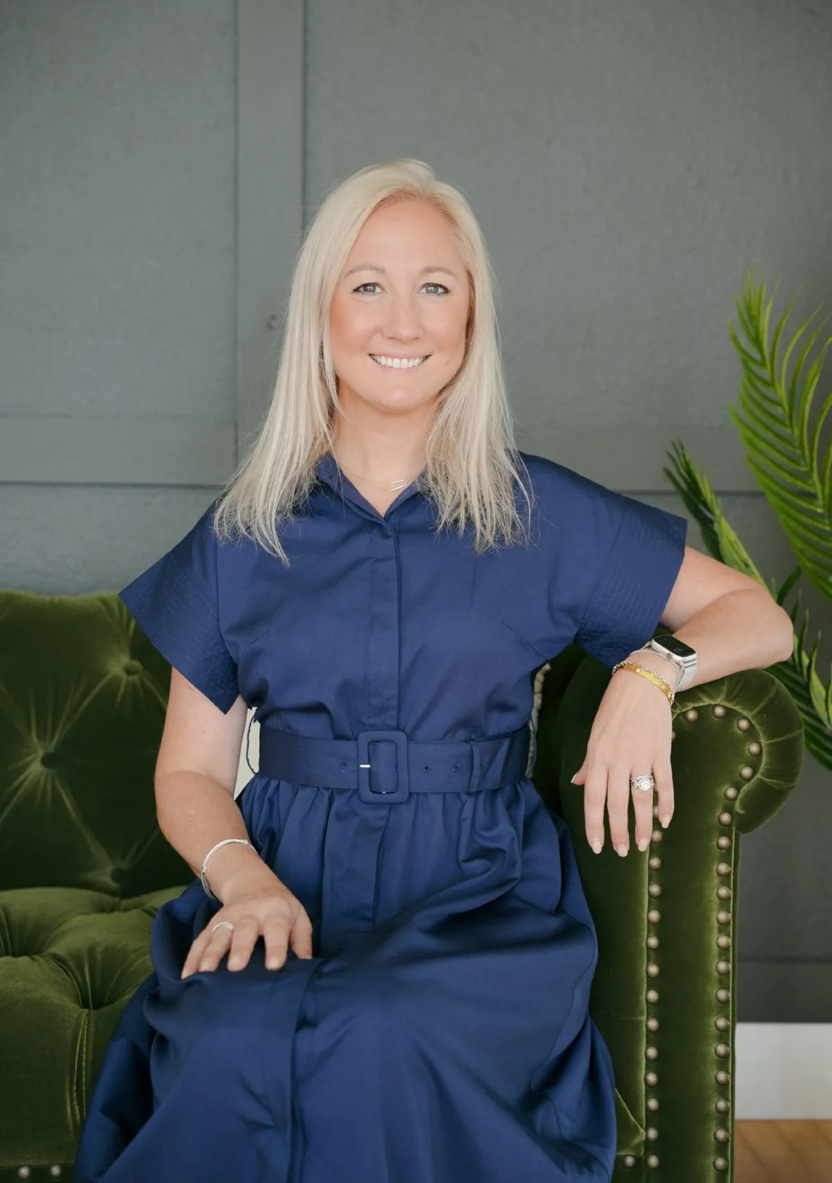 Smiling woman with blonde hair wearing a blue belted dress, sitting on a green velvet couch with studded armrest.