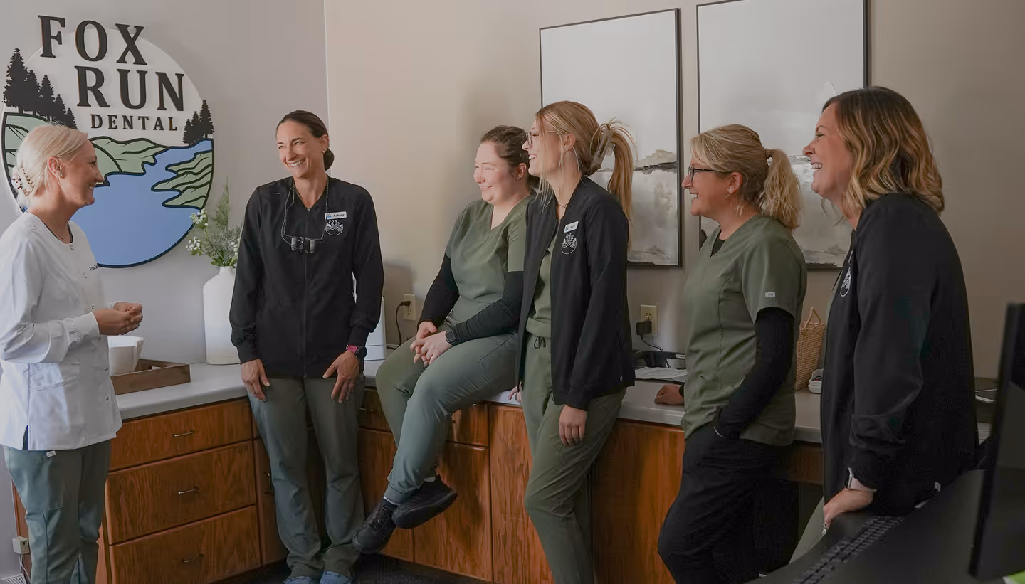 Group of six female dental professionals smiling and talking in a dental office with a Fox Run Dental sign on the wall.