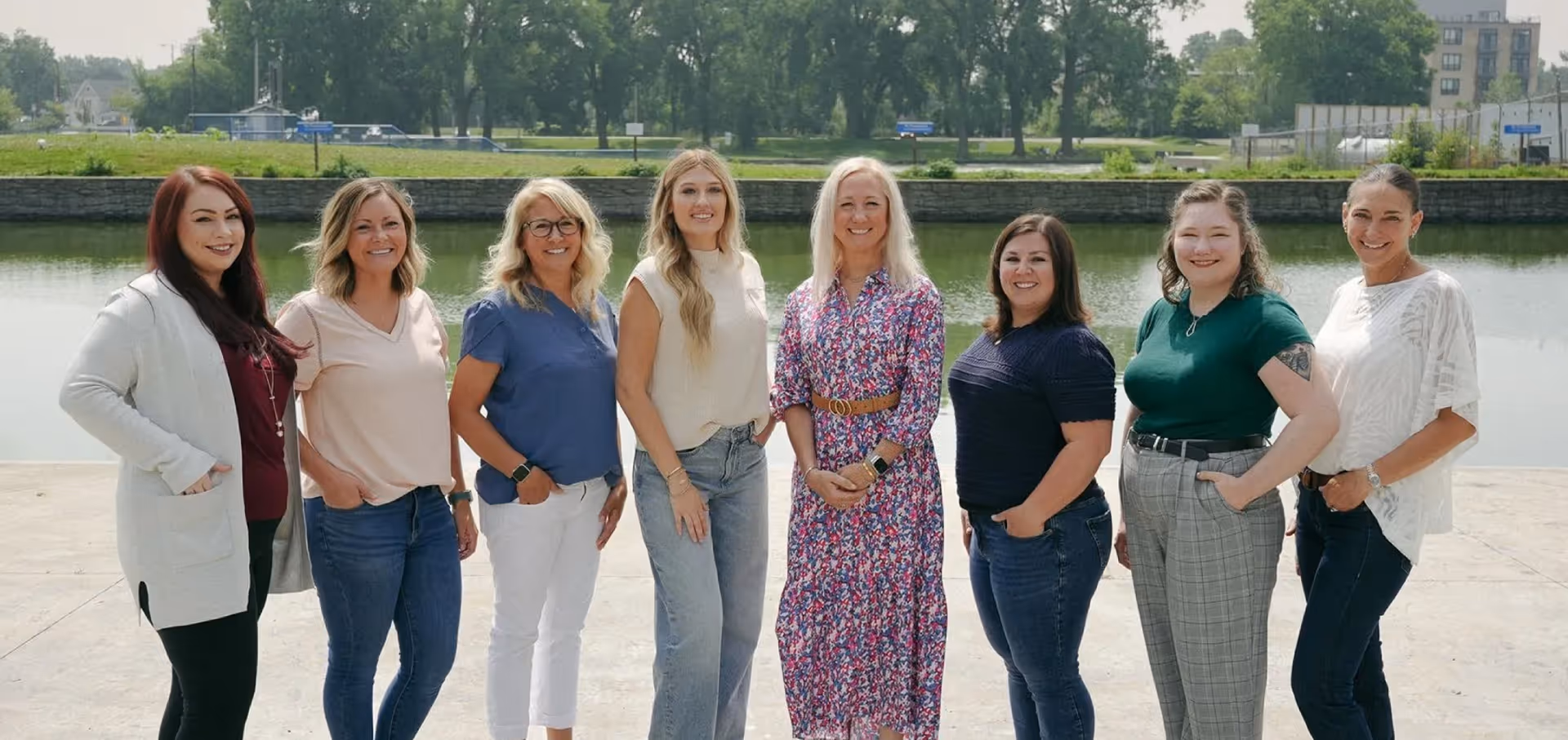 Eight women standing side by side outdoors near a calm river with trees and buildings in the background, all smiling at the camera.