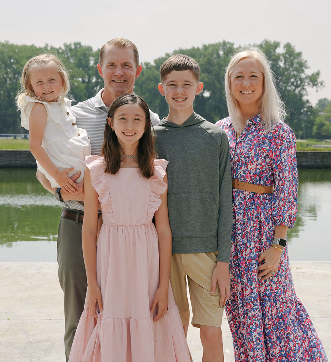 Family of five standing outdoors by a body of water with trees in the background, smiling at the camera.