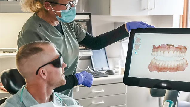 Dental professional wearing a mask and gloves shows a digital 3D dental scan on a screen to a male patient wearing protective glasses.