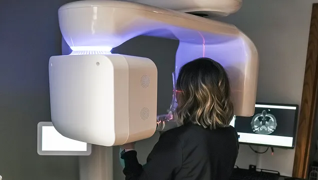 Person undergoing a dental cone beam CT scan with a large white imaging device and brain scan displayed on a computer monitor.