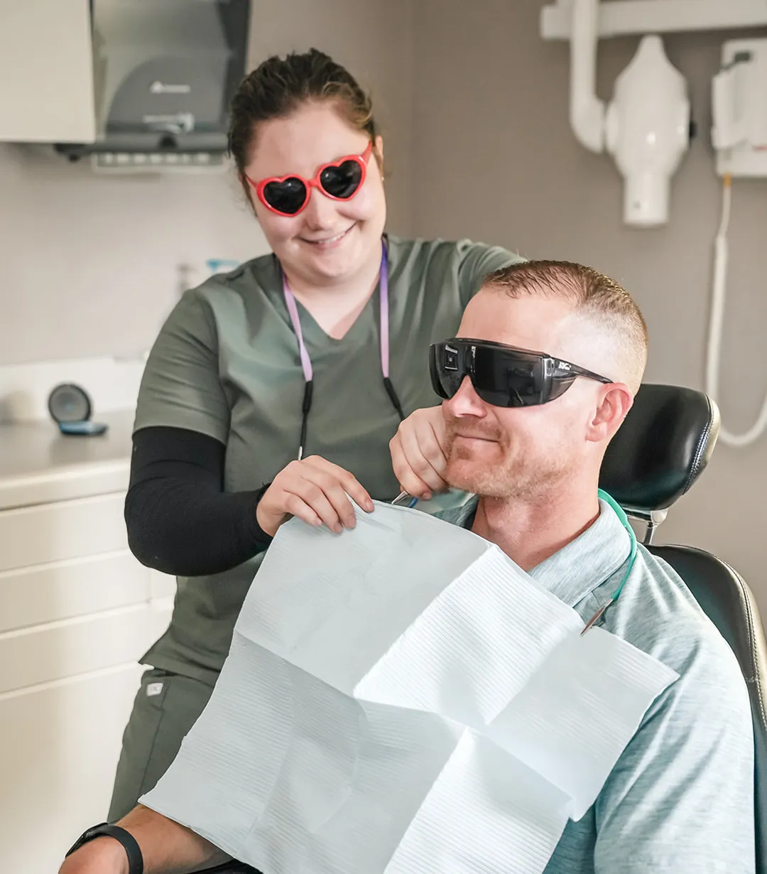 Smiling dental hygienist wearing heart-shaped sunglasses fitting a dental bib on a male patient with protective glasses seated in a dental chair.