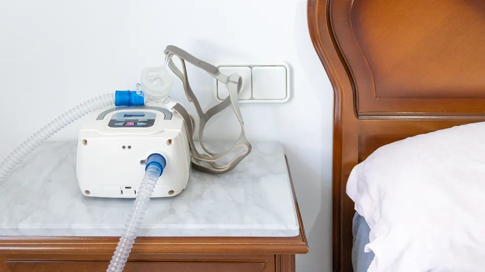 CPAP machine with attached mask on a bedside table next to a wooden bed.