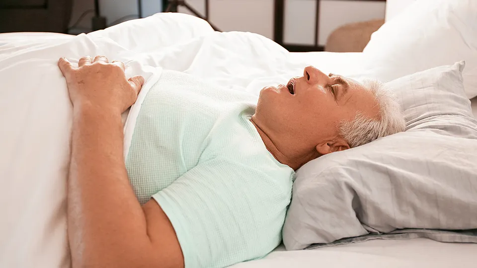 Older man lying on his back asleep in bed with mouth slightly open and hands resting on covered chest.