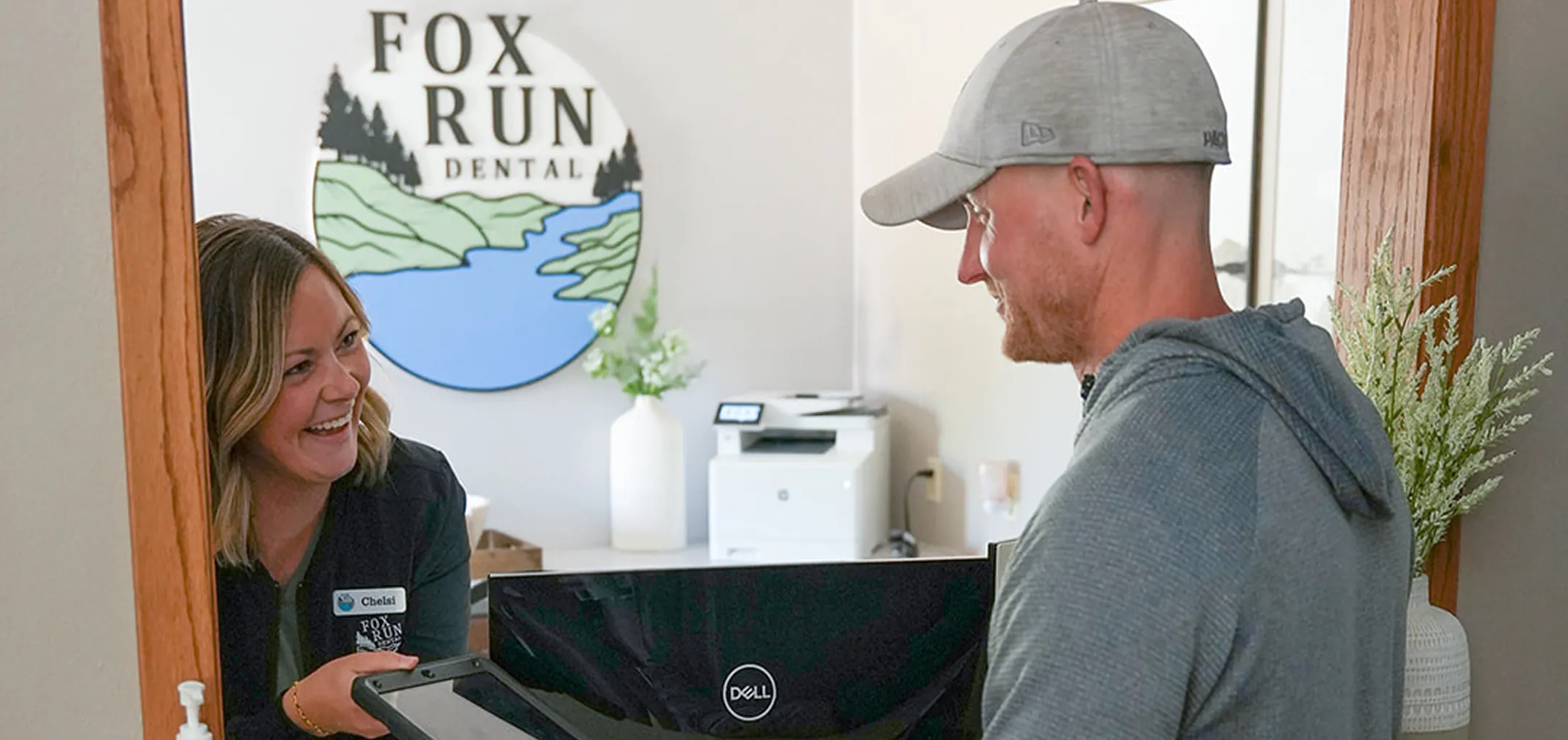 Smiling dental receptionist handing a tablet to a male patient at a clinic counter with Fox Run Dental logo in the background.