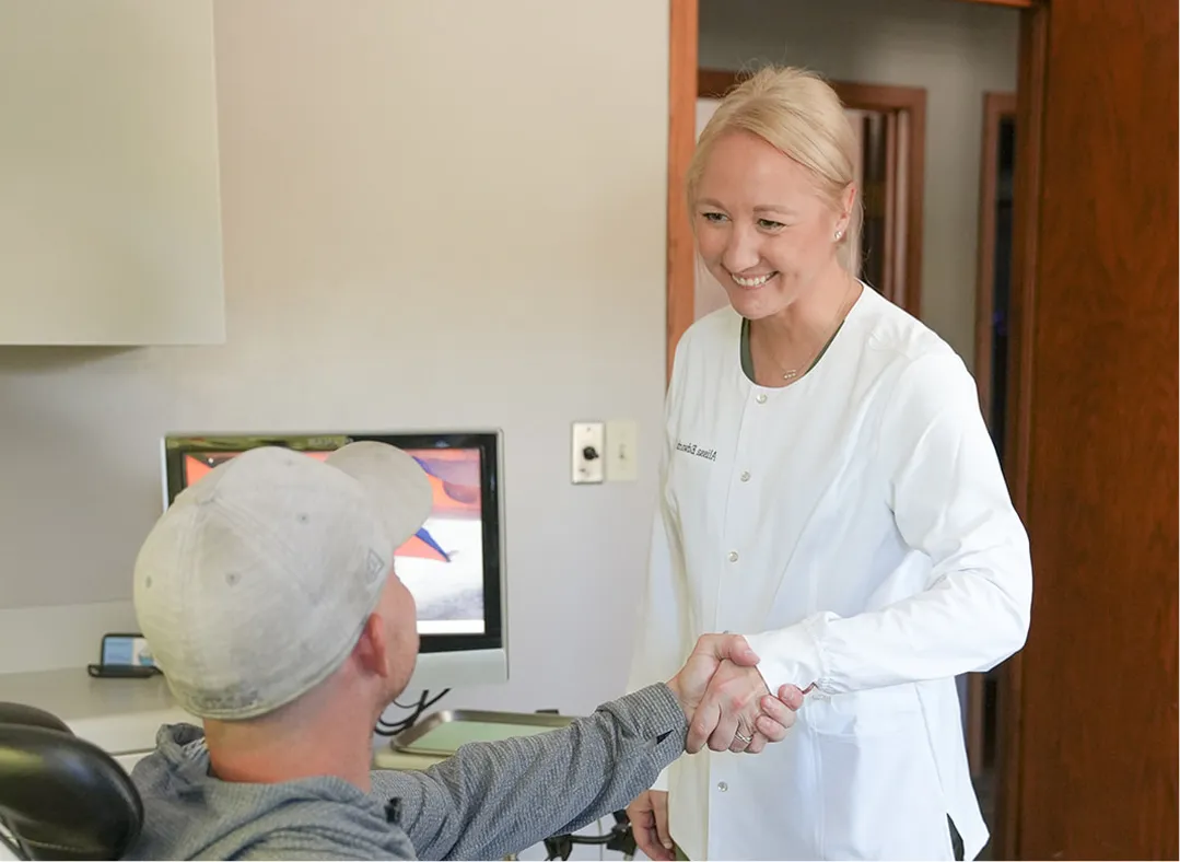 Smiling female healthcare professional shaking hands with a seated male patient wearing a gray cap and sweatshirt in a clinical office.