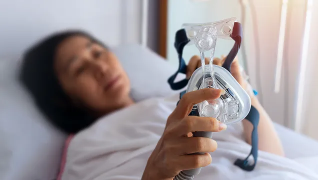 Person lying in bed holding a clear CPAP mask used for sleep apnea treatment.