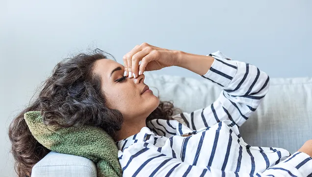 Woman lying on a sofa with eyes closed and pinching the bridge of her nose, appearing to have a headache.