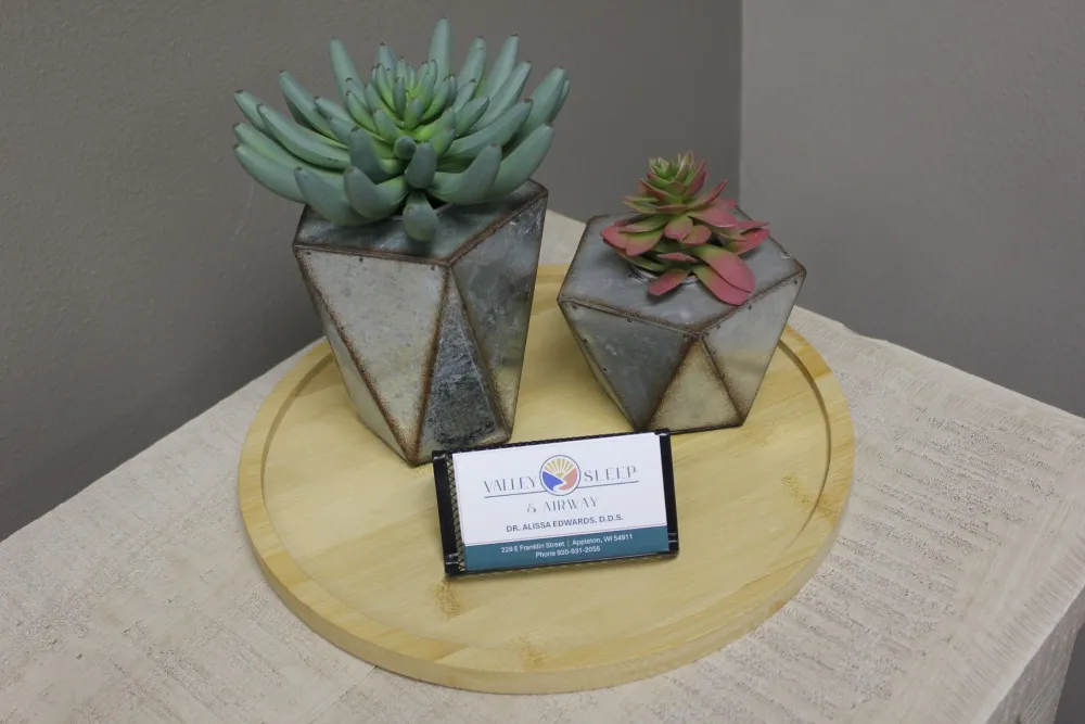 Two geometric metal planters with succulents on a round wooden tray atop a textured beige cloth.