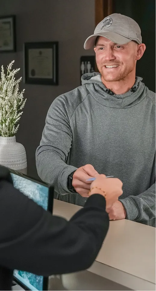Smiling man in a gray hoodie and cap handing a card over a reception counter.