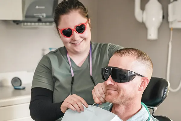 Dentist wearing heart-shaped red sunglasses smiling while a male patient with protective glasses sits in a dental chair.