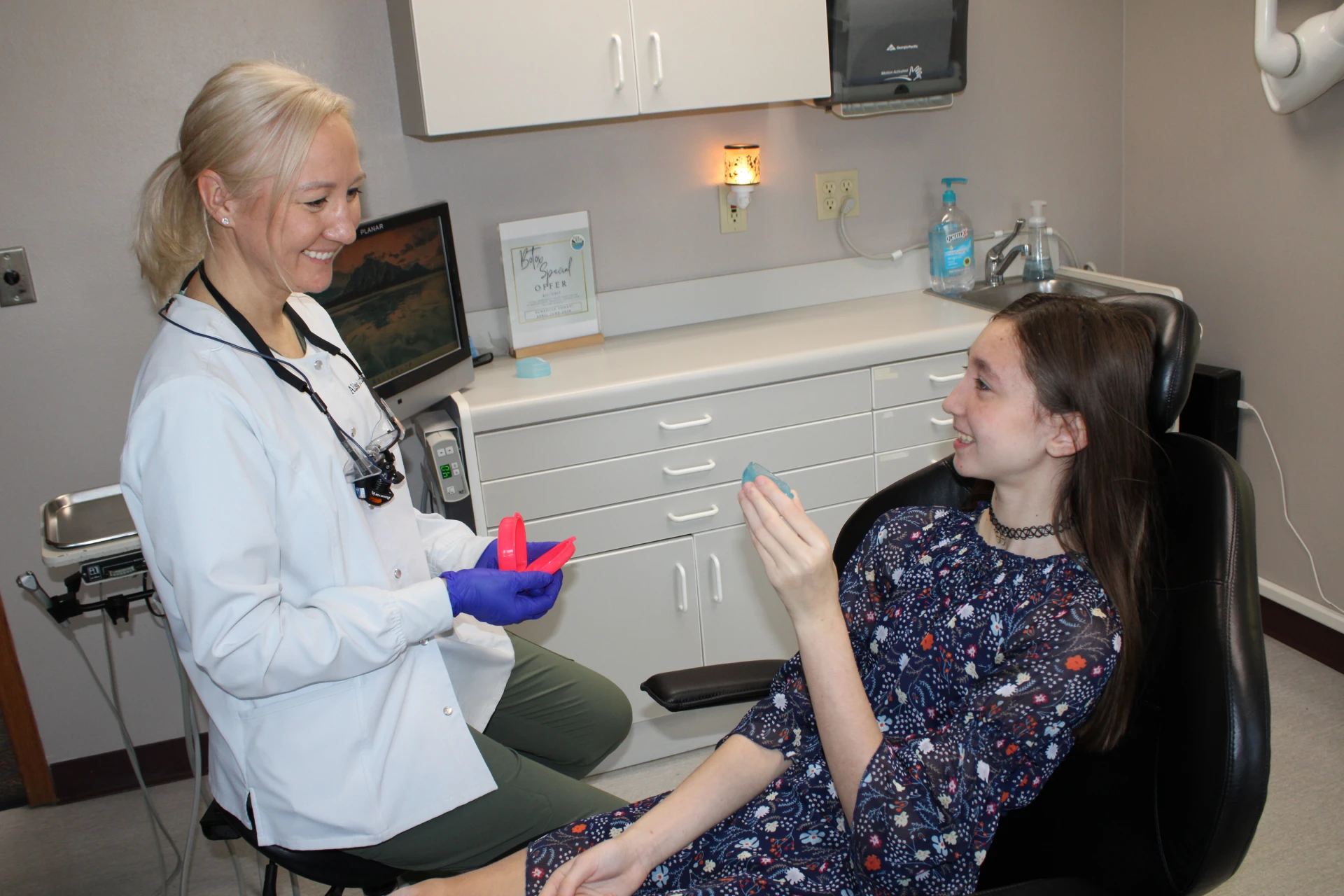 Smiling female dentist in white coat and purple gloves showing dental retainer cases to a young girl sitting in a dental chair holding a clear retainer.
