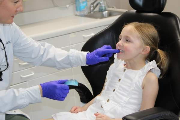 Dentist wearing purple gloves fitting a blue dental guard into a smiling young girl's mouth in a dental office.