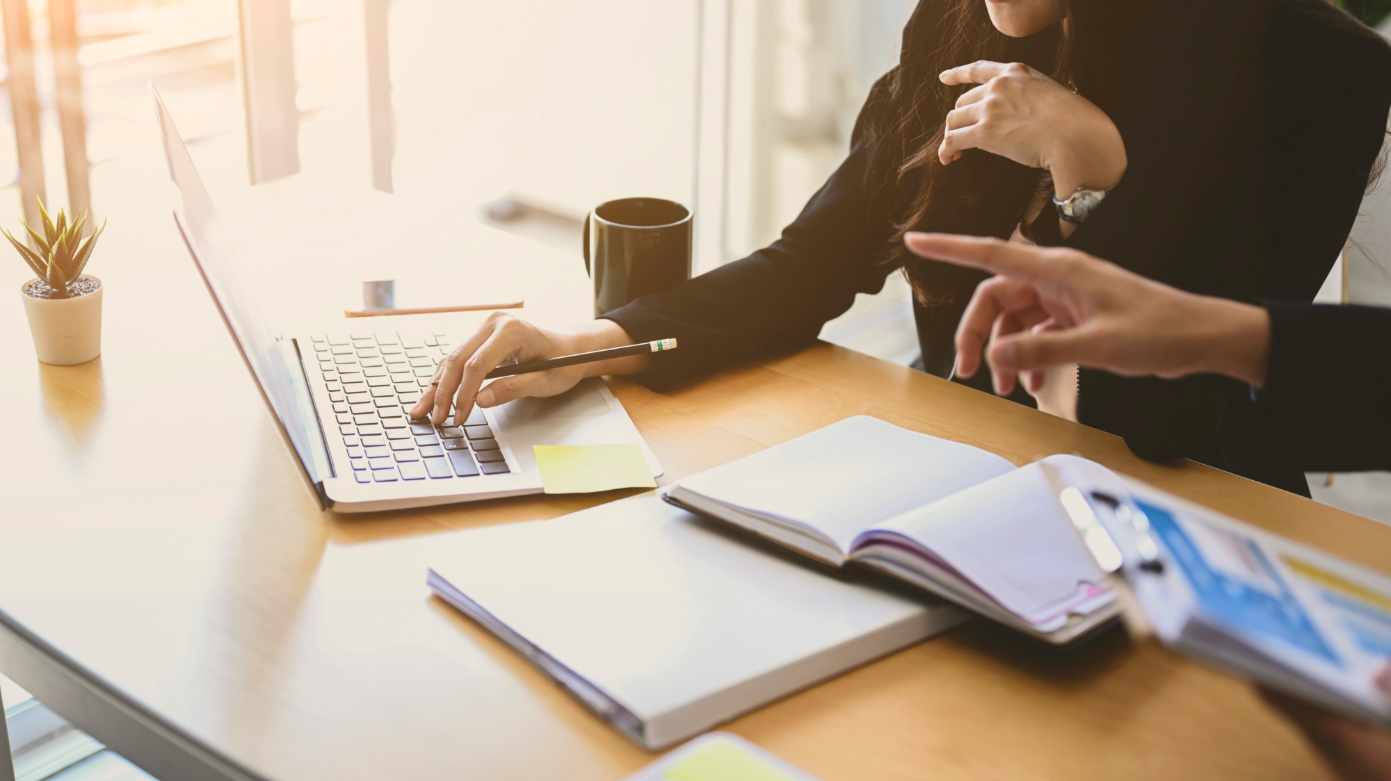 Two people working together at a table, one on a laptop, and one pointing to the laptop.
