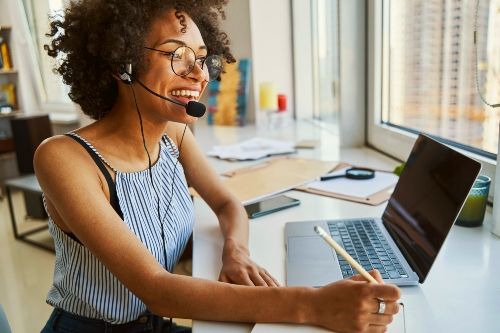 Women sitting at a desk with a headset on taking a phone call while writing with a pencil in front of a laptop.