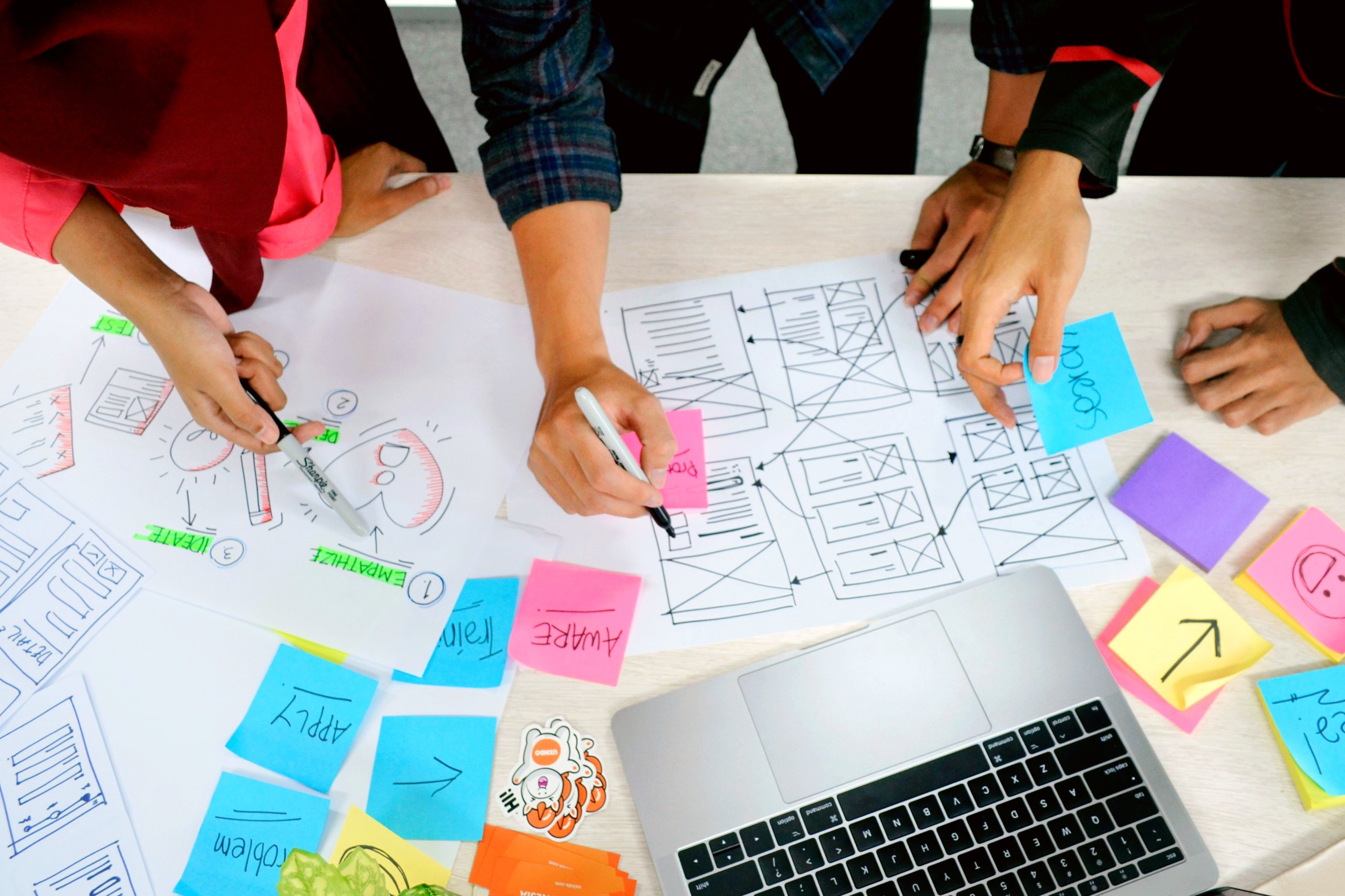 Overhead view of three people's hands working together with papers and a laptop. Drawing diagrams and taking notes.