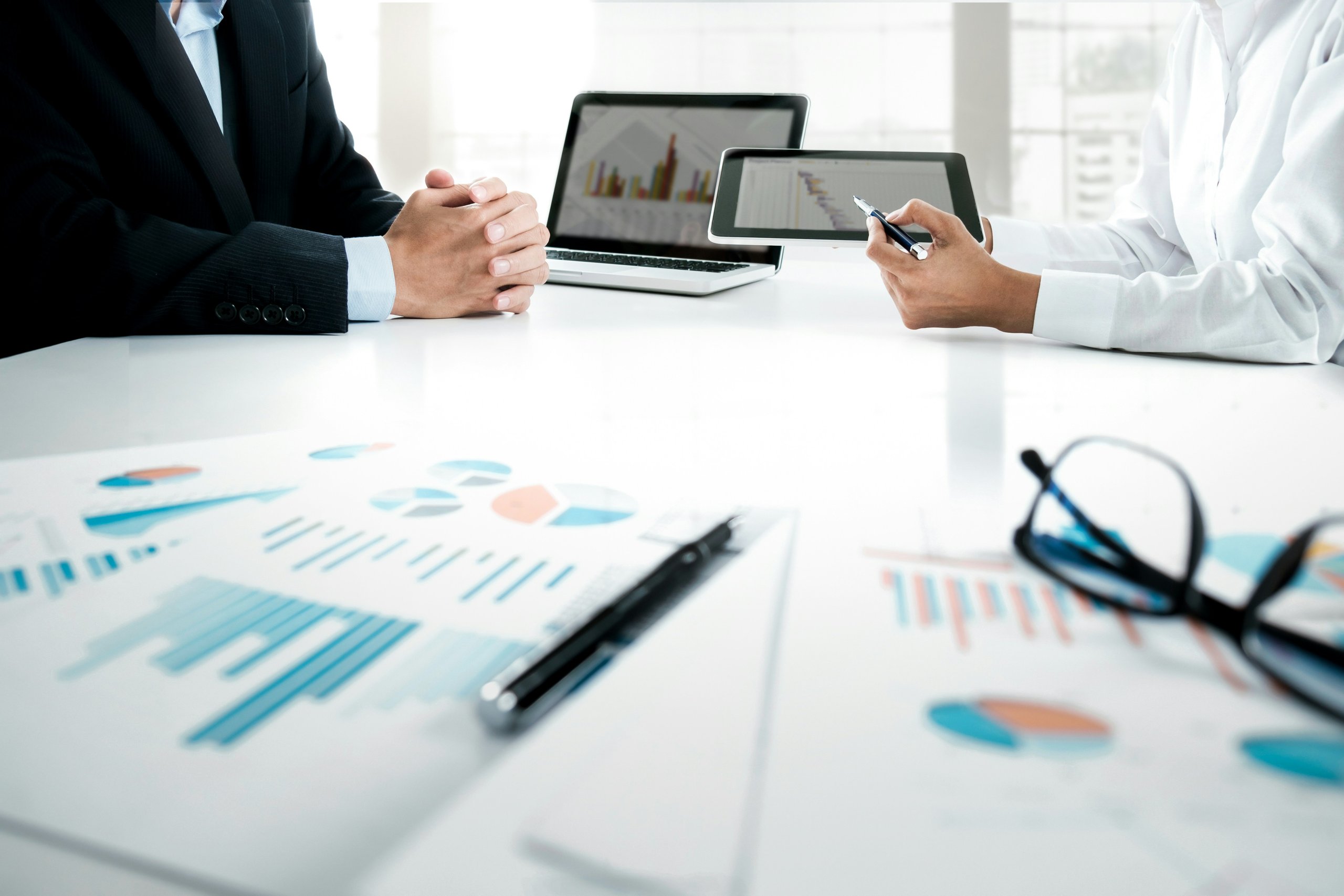Hands of two people working together at a table that has papers of diagrams and graphs thrown about.  With two laptops displaying graphs.