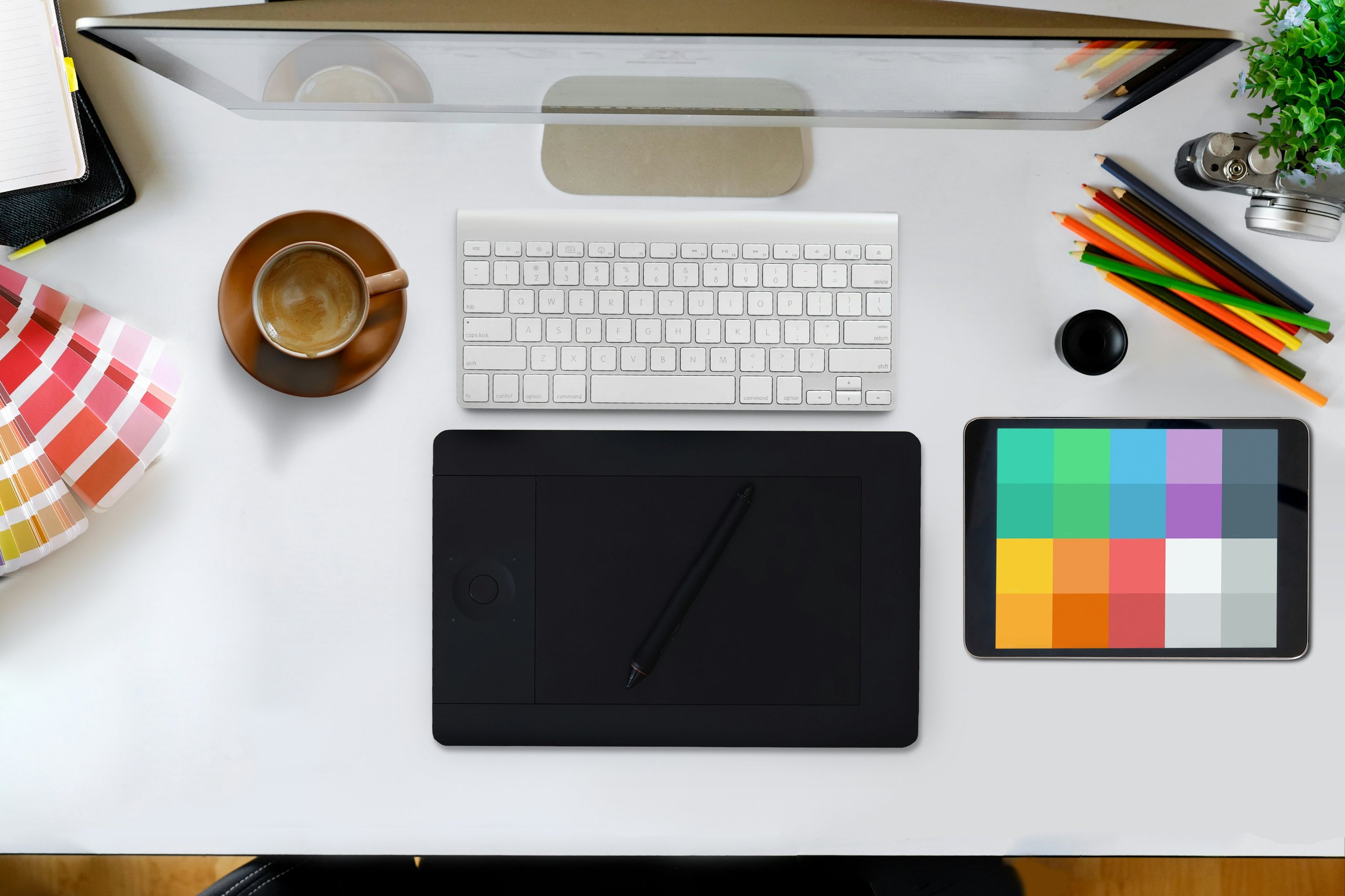 Overhead view of a desk with a computer monitor a cup of coffee and a color swatch and coloring pencils.