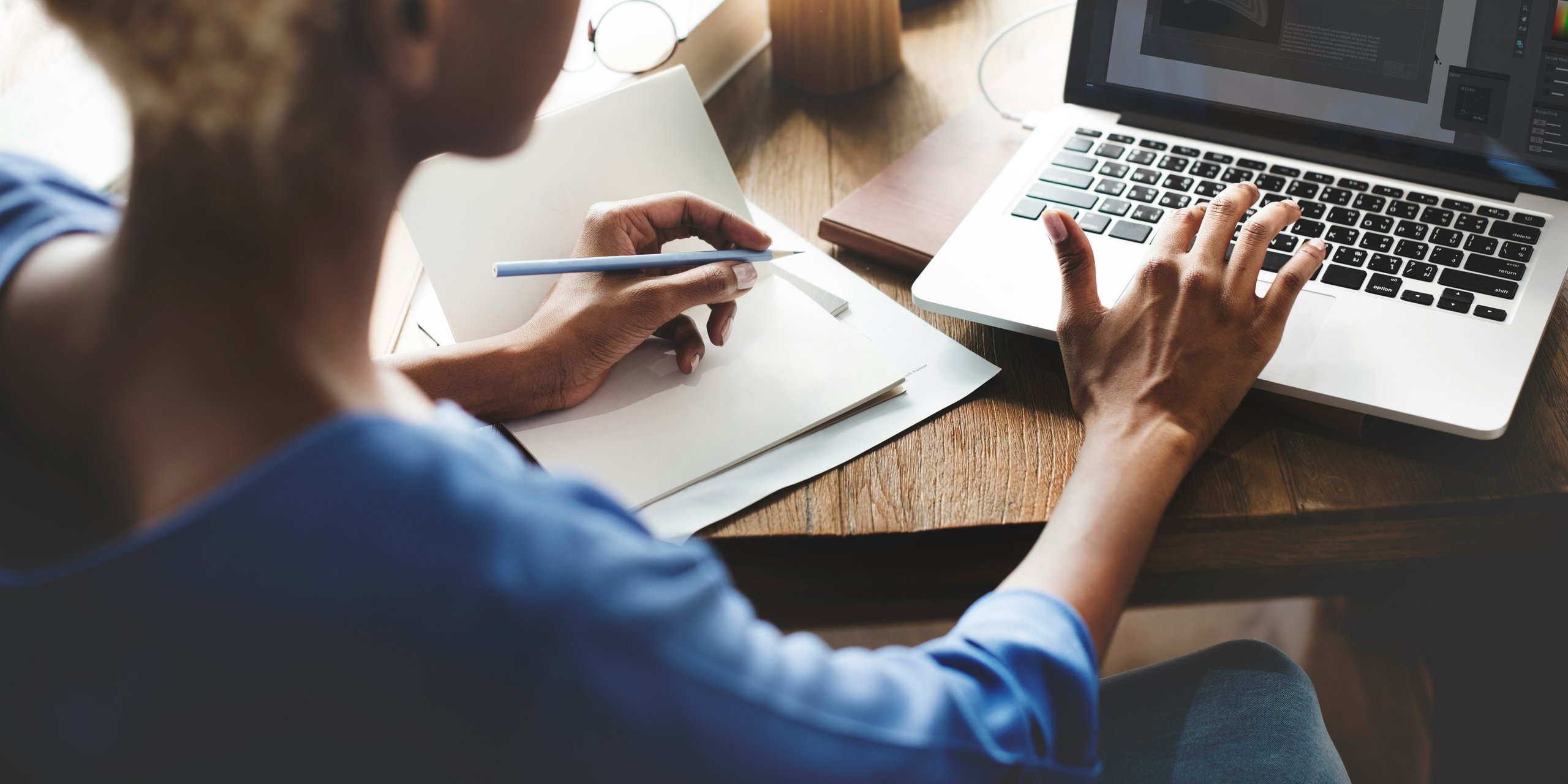 Over the shoulder of a women taking notes in a notepad and typing on a laptop.