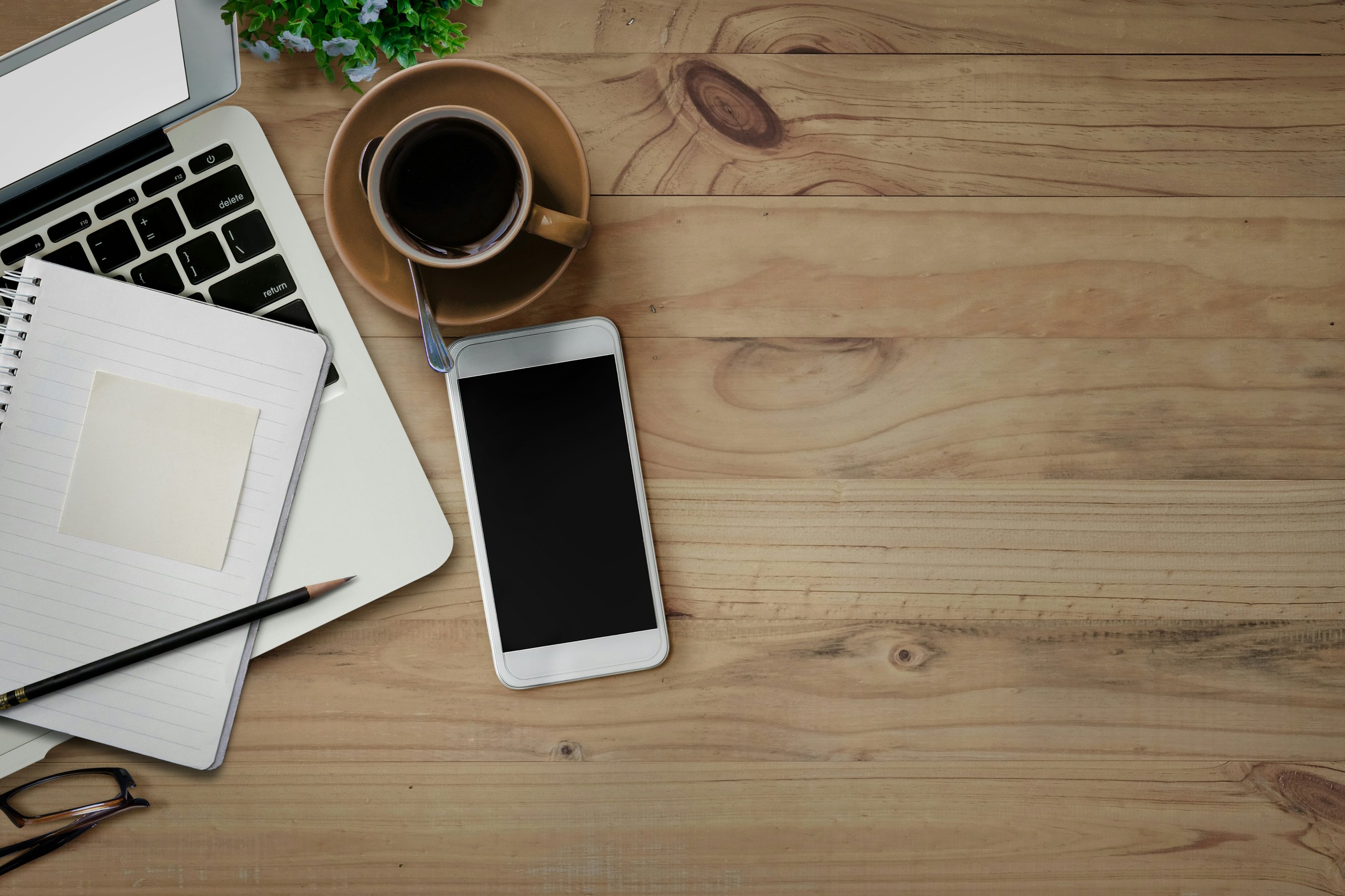 Overhead view of a desk with an open laptop that has a notepad on the keyboard, next to a cellphone and cup of coffee.