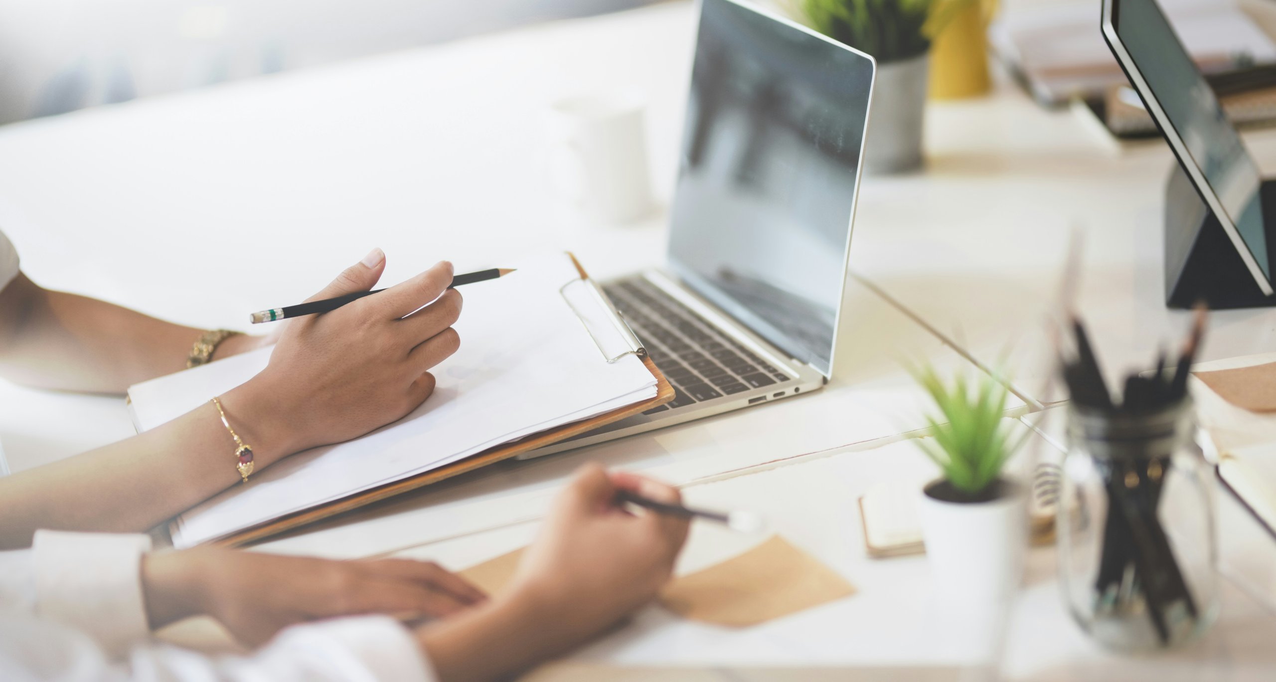 Two people's hands holding a pen and clipboard next to a laptop.