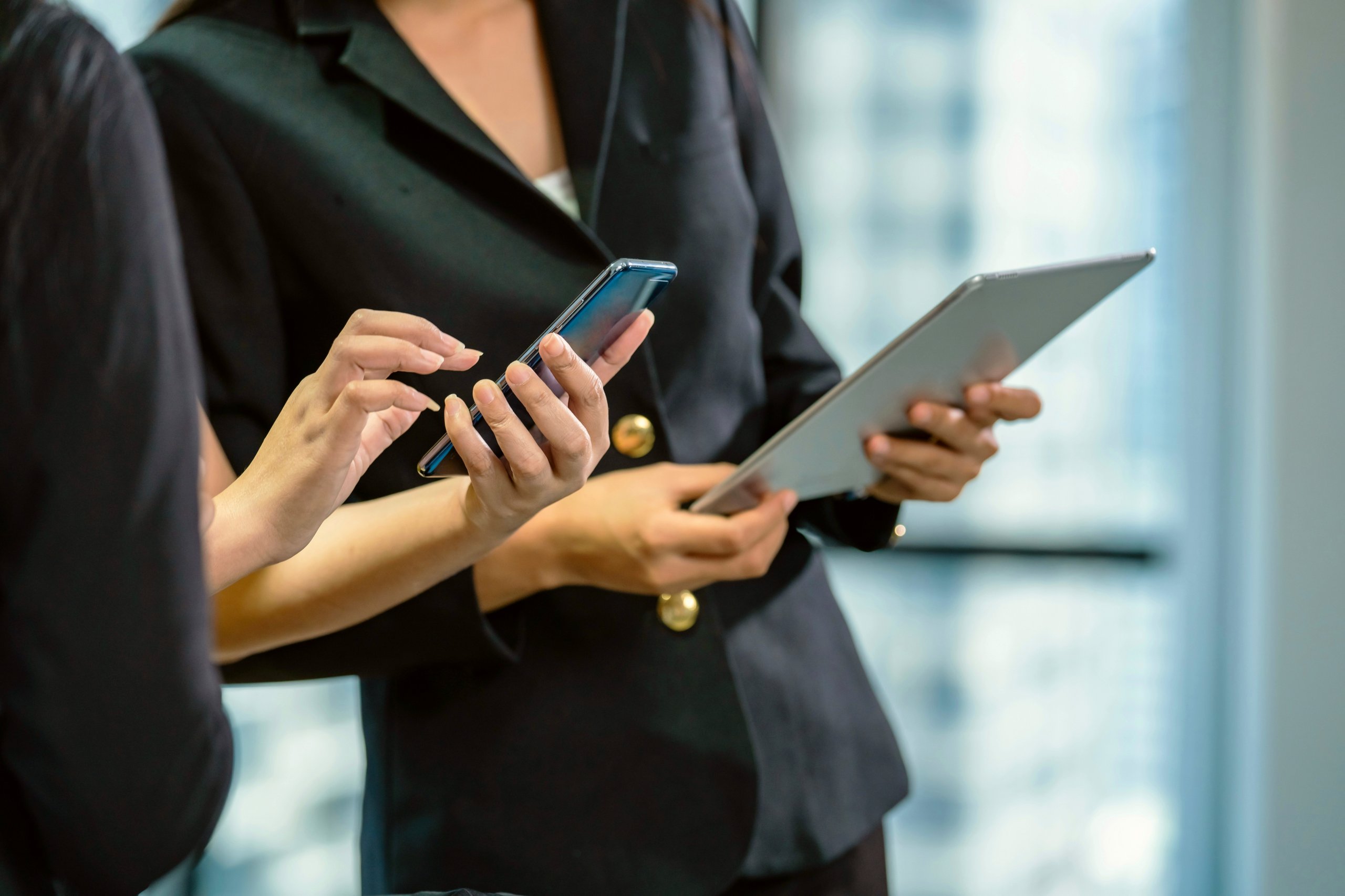 A women holding a cell phone standing beside a women holding a tablet.