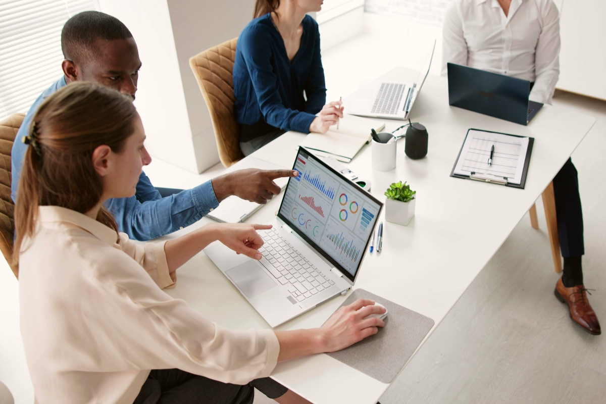 A man and a women working together reviewing a graph on a laptop in a small meeting room among two others.  