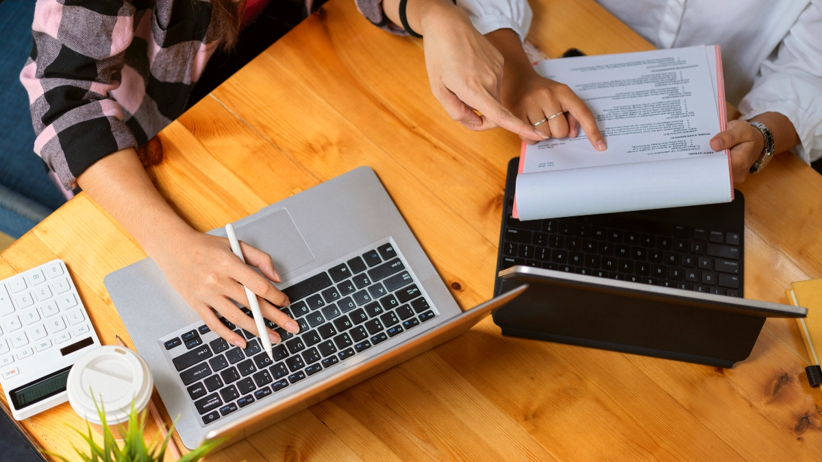 Hands of two women working together reviewing a document and using a laptop.