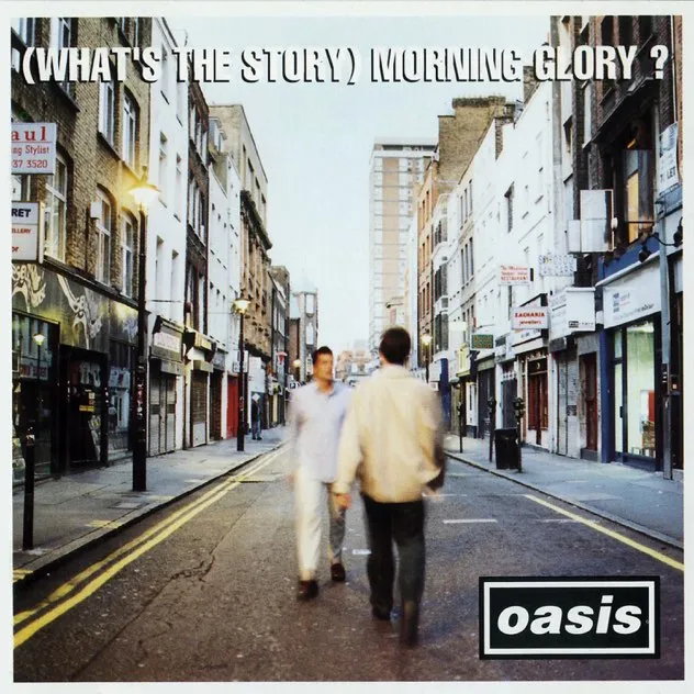 Blurred image of two men walking towards each other on an empty city street with buildings and lampposts on each side, text at the top reads '(WHAT'S THE STORY) MORNING GLORY?' and a black box with 'oasis' in white letters at the bottom right.