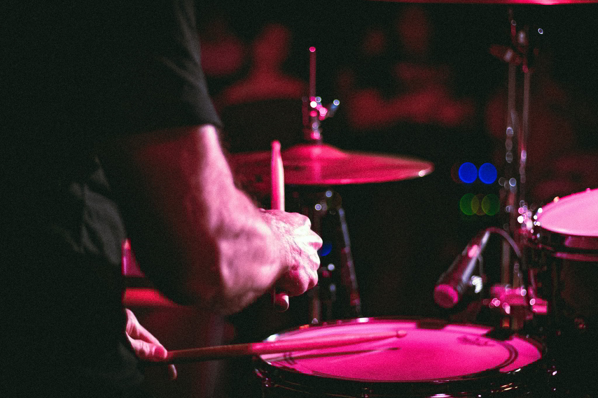 Close-up of a drummer's hands playing a drum set under pink stage lighting. bandkaroke.live