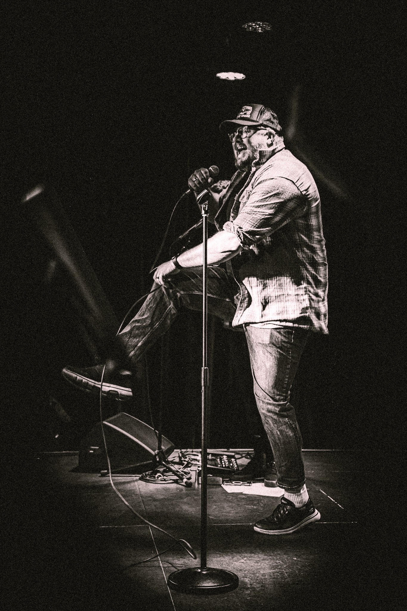 Black and white image of a man wearing a cap and glasses performing energetically on stage with a microphone, lifting one leg.