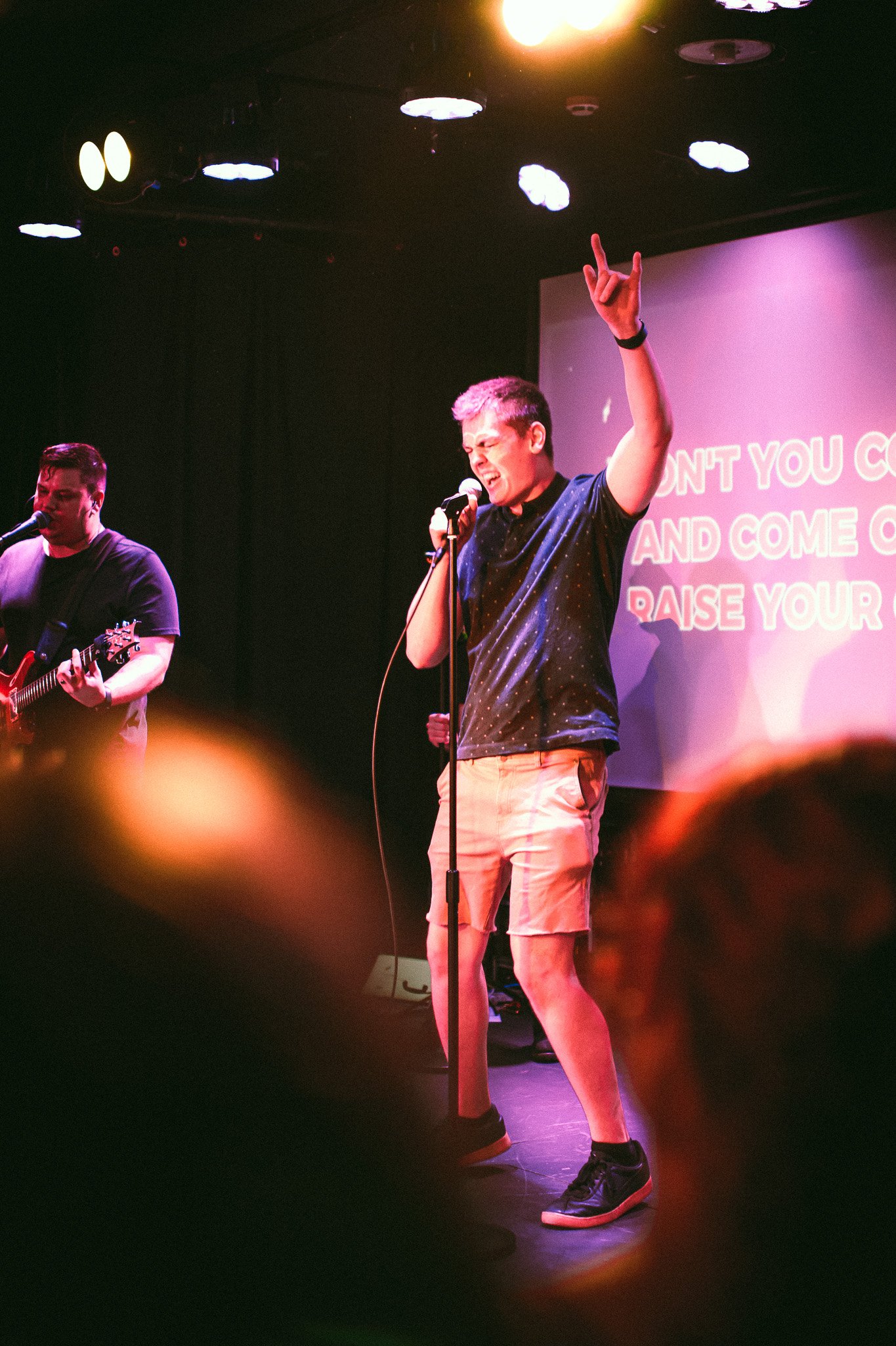 Man singing passionately into a microphone on stage with one hand raised, accompanied by guitarist, under stage lights. bandkaroke.live