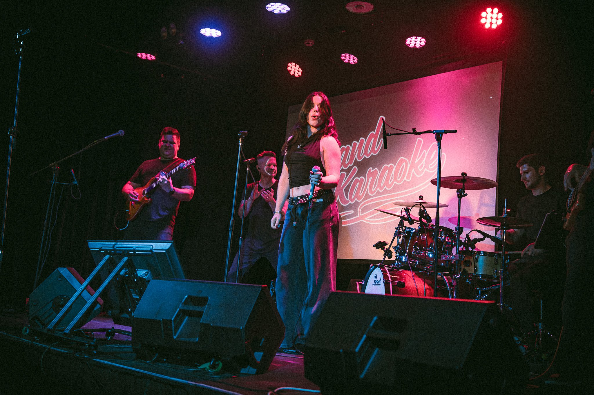 A female singer holding a microphone on stage with a band playing guitar, drums, and clapping beside her under colorful stage lights. bandkaroke.live