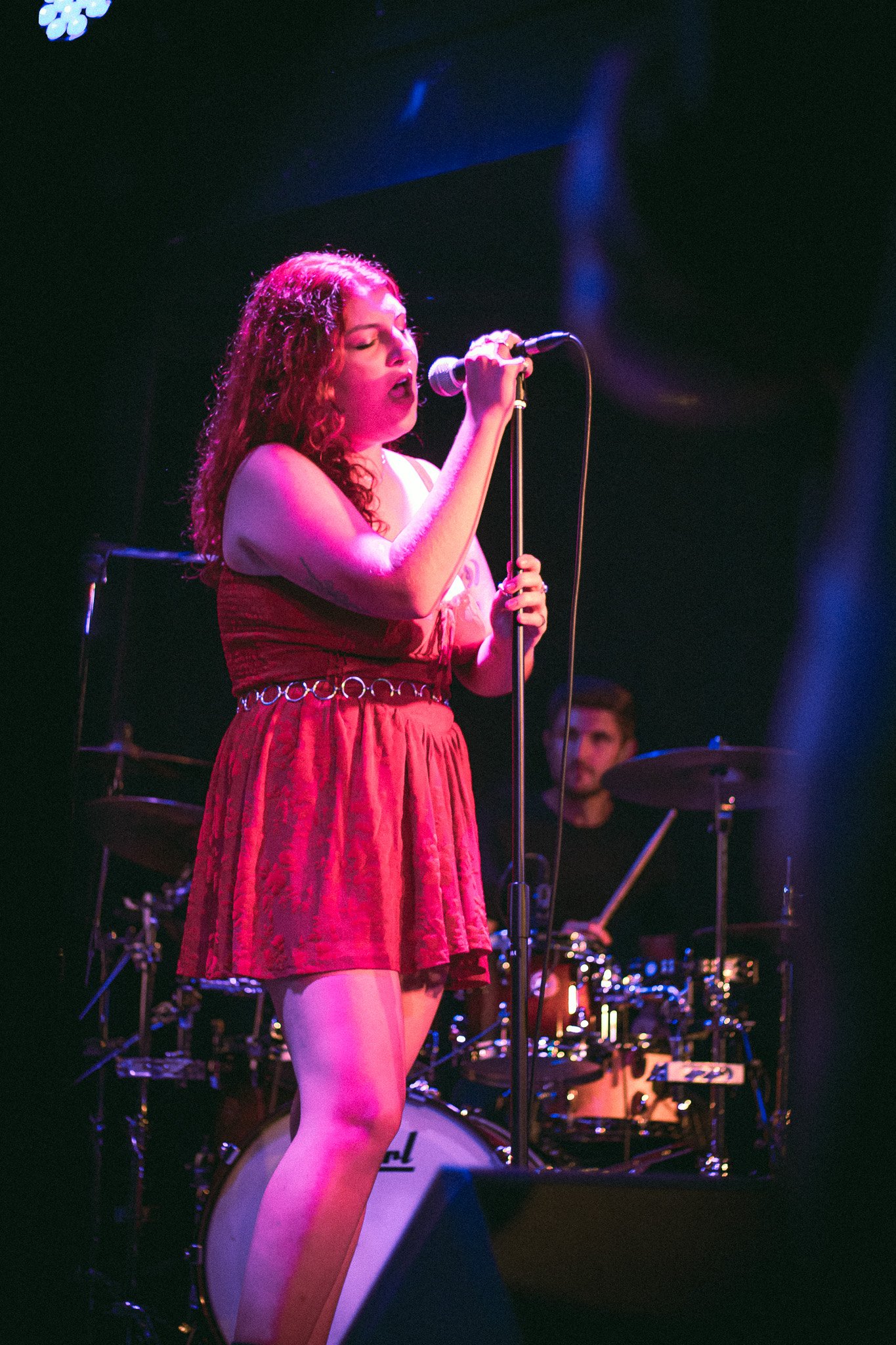 Female singer in a red dress performing on stage with a drummer in the background. bandkaroke.live