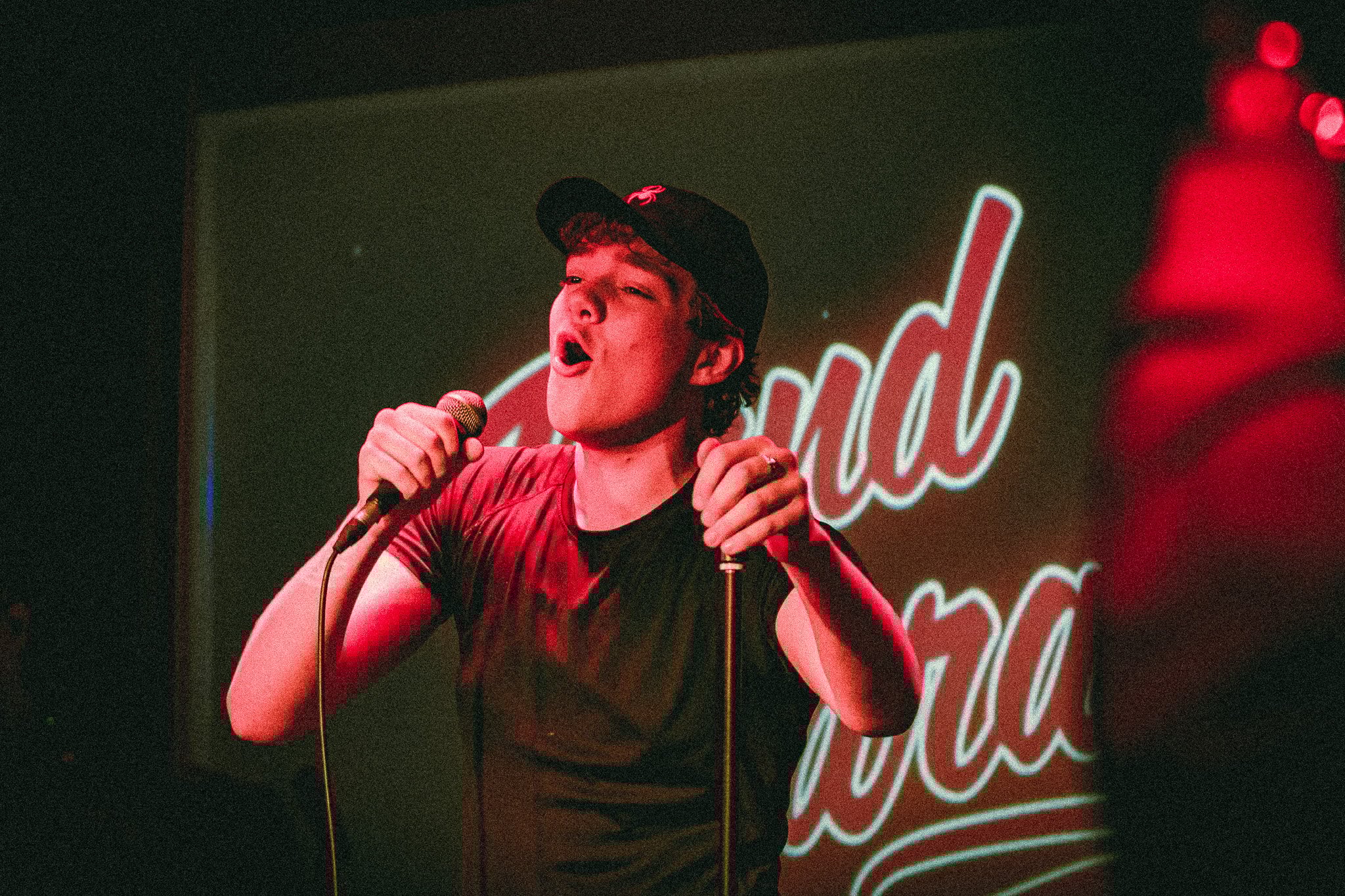 Young man wearing a black cap and shirt singing passionately into a microphone on stage with red lighting and partial blurred text in the background. bandkaroke.live
