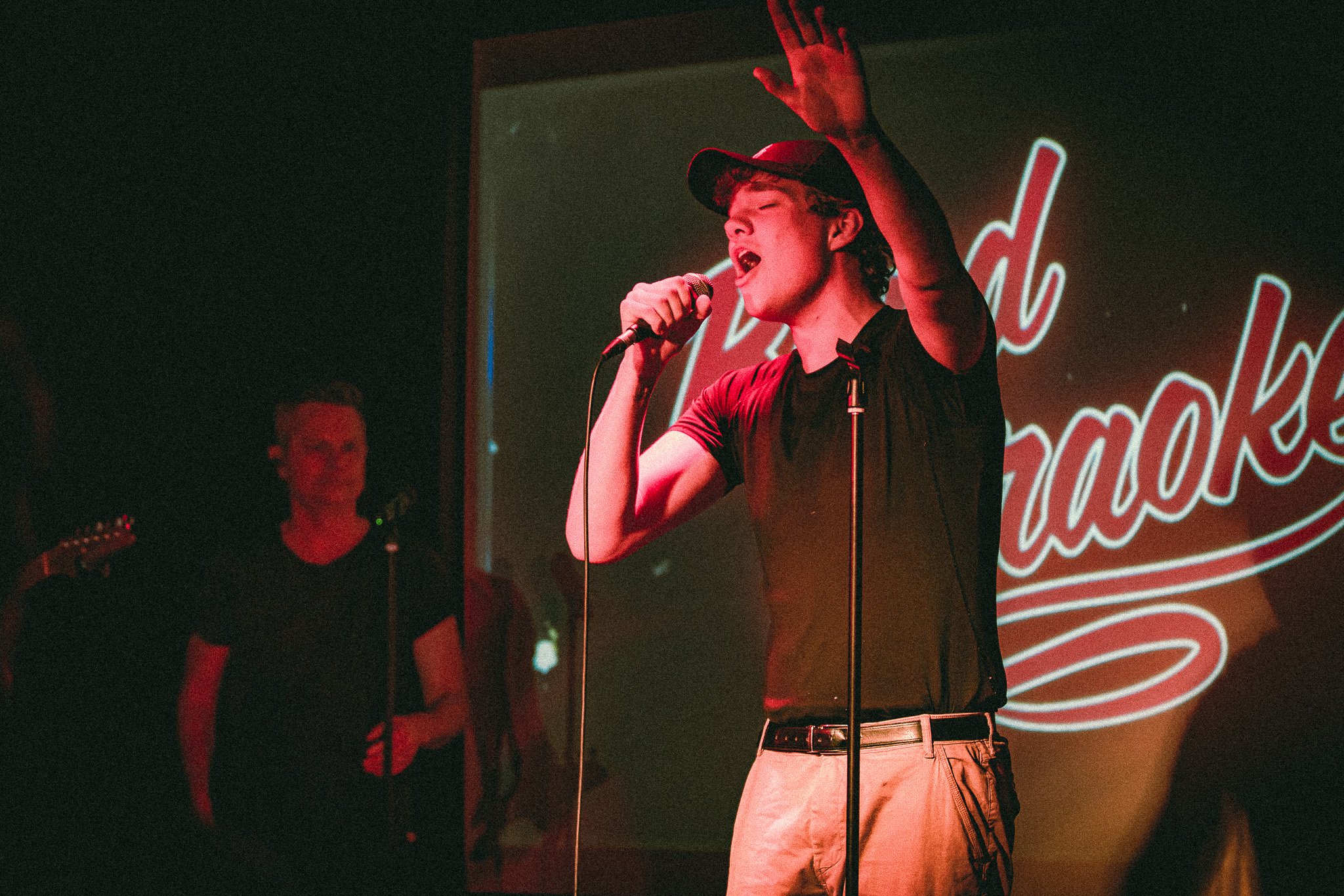 Young man wearing a cap singing passionately into a microphone on stage at a karaoke event. bandkaroke.live
