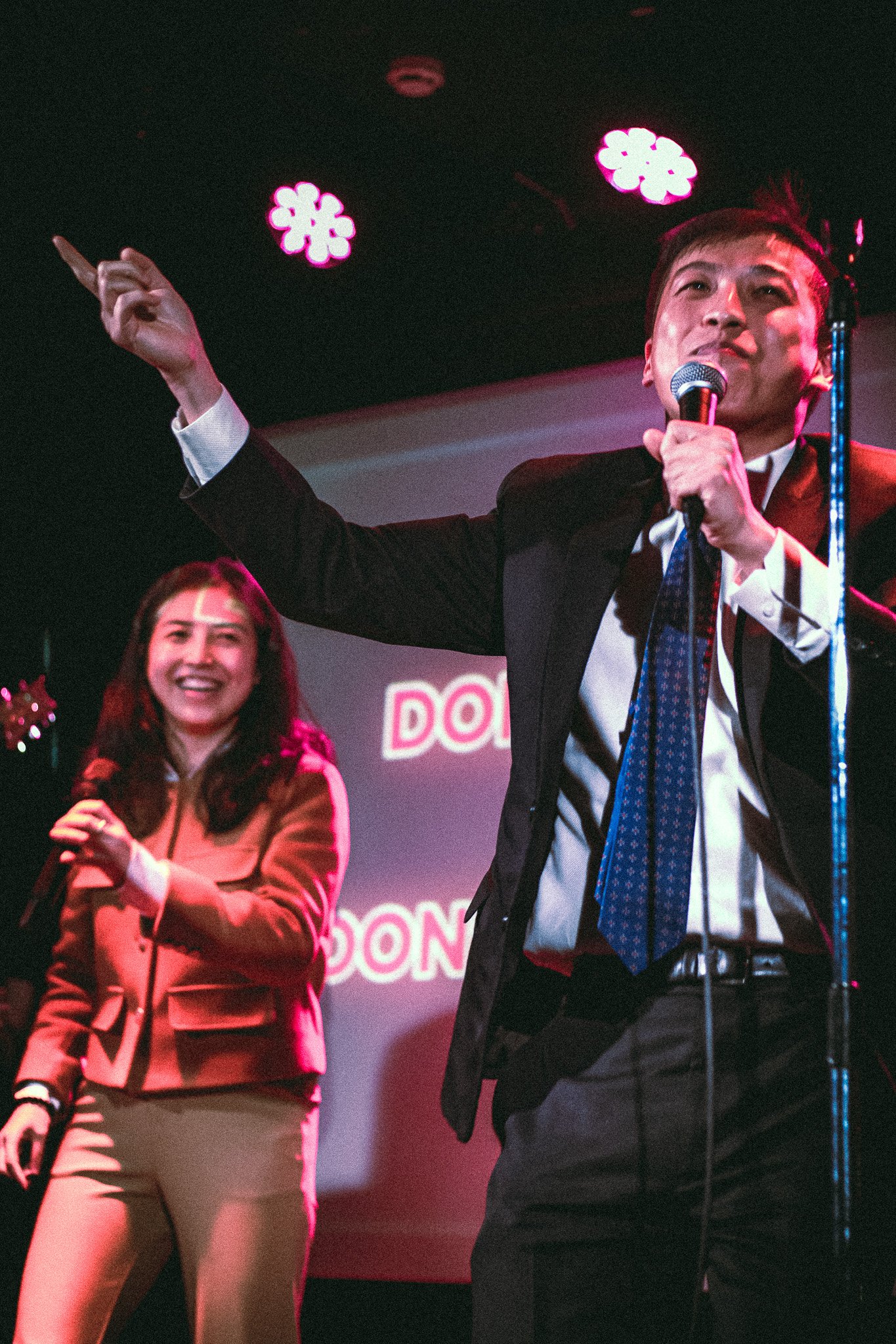 Man in a suit and tie speaking into a microphone and pointing, with a smiling woman holding a microphone beside him on stage under pink lights. bandkaroke.live