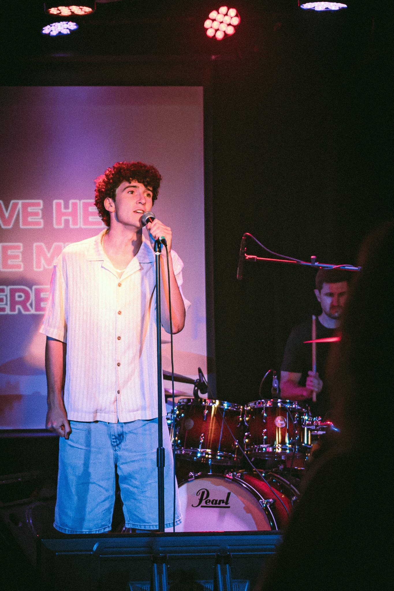 Young man singing into a microphone on stage with a drummer playing behind him. bandkaroke.live