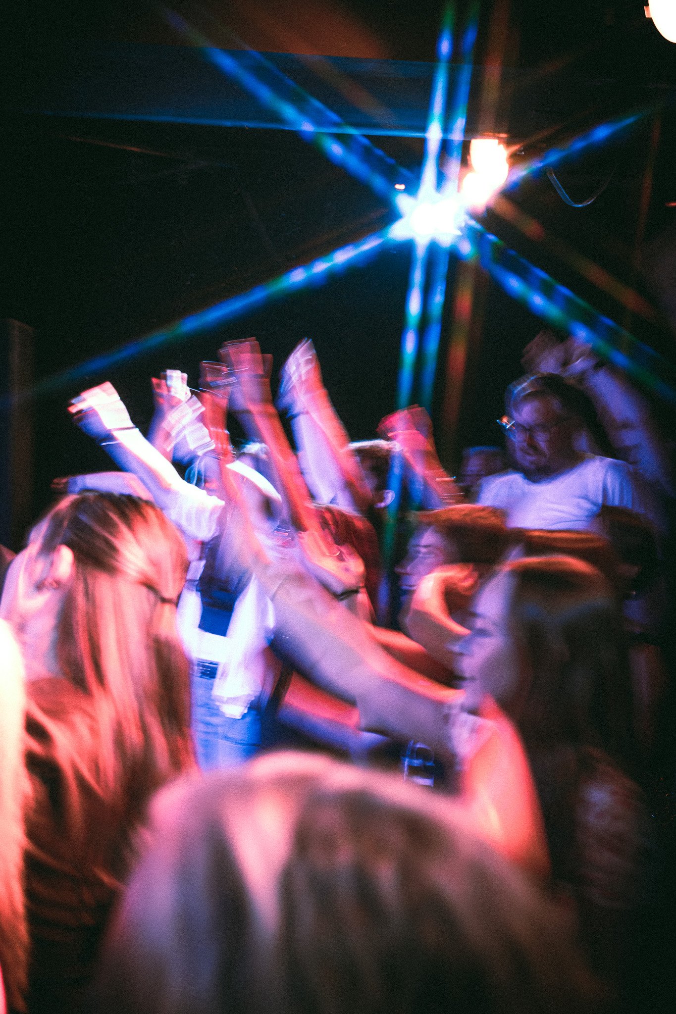Blurred crowd dancing with raised hands under colorful stage lights at a party or concert. bandkaroke.live